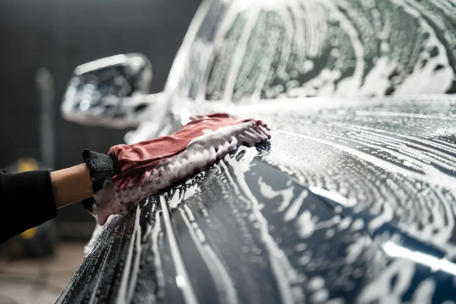 Hand with a red wash mitt wiping suds across a car hood and windshield during a professional detail